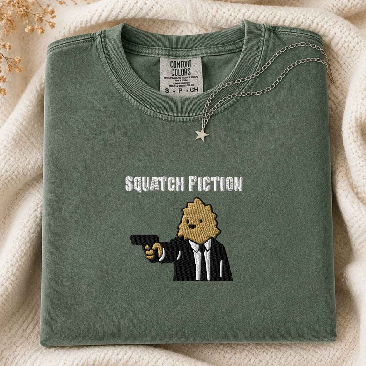 A folded sage green Comfort Colors t-shirt with a graphic of a bigfoot in a suit holding a gun and the text 'SQUATCH FICTION' embroidered on the front. A gold star pendant necklace is draped over the collar, set against a cozy cream knit blanket background.