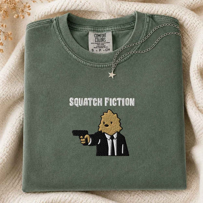 A folded sage green Comfort Colors t-shirt with a graphic of a bigfoot in a suit holding a gun and the text 'SQUATCH FICTION' embroidered on the front. A gold star pendant necklace is draped over the collar, set against a cozy cream knit blanket background.