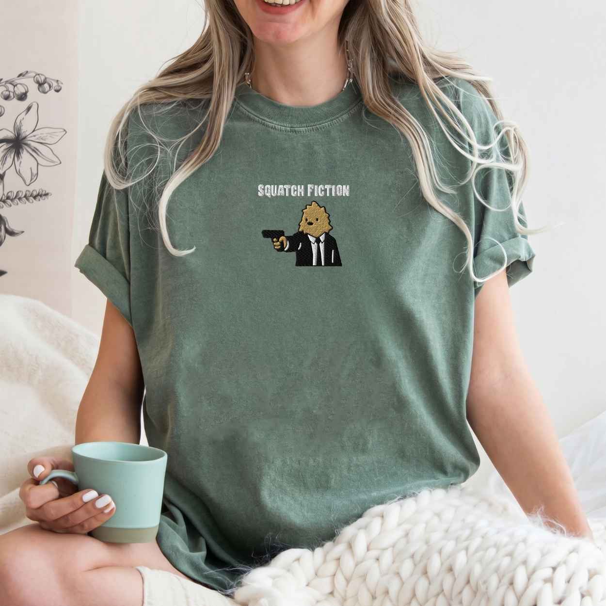 A woman wearing a sage green graphic t-shirt with a cartoon bigfoot and the text 'SQUATCH FICTION' printed on the chest. She is holding a mint green mug while sitting on a bed with a chunky knit blanket.