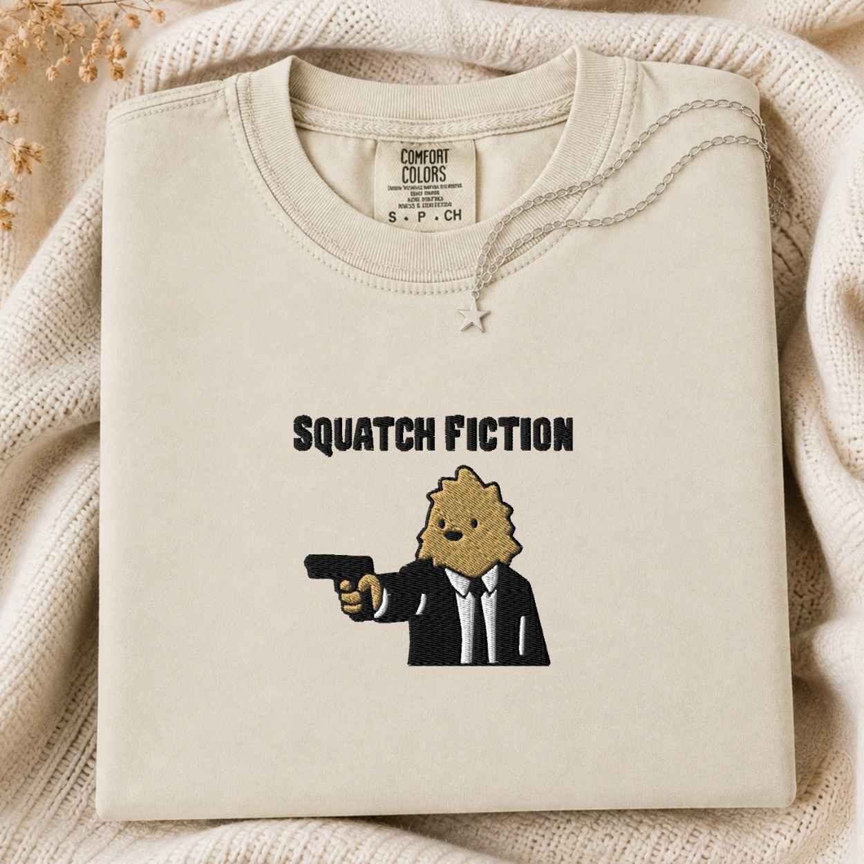 A folded cream-colored tshirt with a graphic print of a bigfoot in a suit holding a gun, with the text 'SQUATCH FICTION' above it. The tshirt is displayed on a cozy, textured cream knit blanket with dried flowers in the corner.