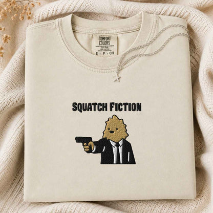 A folded cream-colored tshirt with a graphic print of a bigfoot in a suit holding a gun, with the text 'SQUATCH FICTION' above it. The tshirt is displayed on a cozy, textured cream knit blanket with dried flowers in the corner.