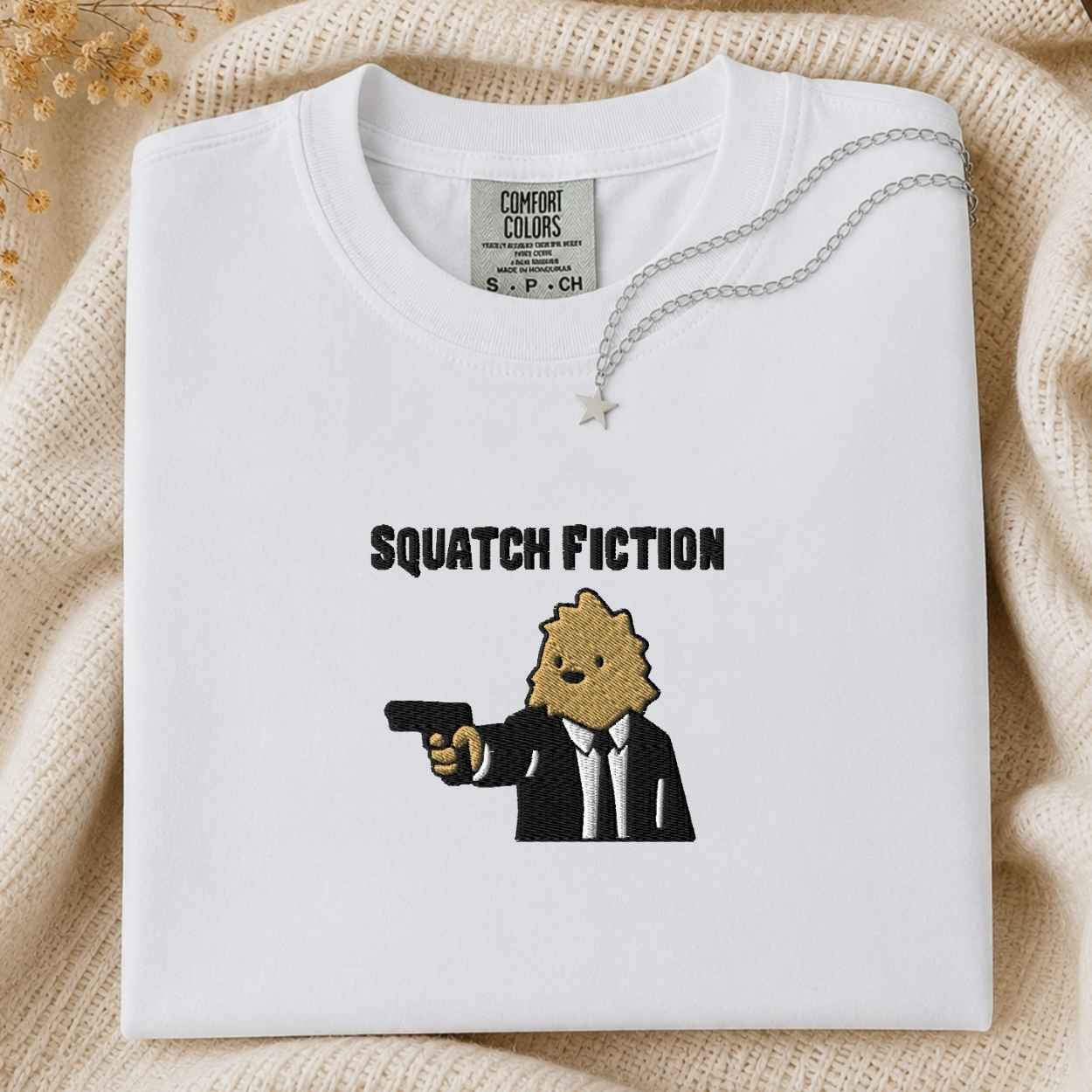 A folded white Comfort Colors t-shirt with a graphic print of a cartoon bigfoot in a black suit holding a gun and the text is SQUATCH FICTION, next to a silver necklace, laid flat on a beige knit blanket.