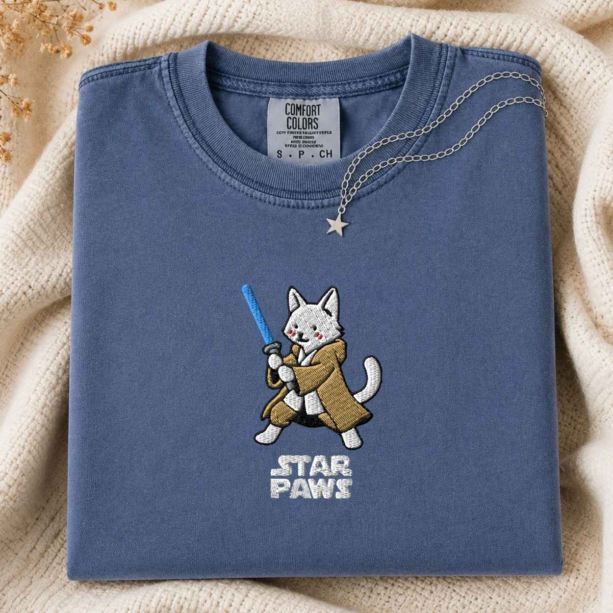 A folded slate blue t-shirt with a cute embroidered white cat character wearing a brown robe and holding a blue laser, with 'STAR PAWS' embroidered below. The shirt is styled on a beige knit blanket with a silver star necklace draped over the collar.