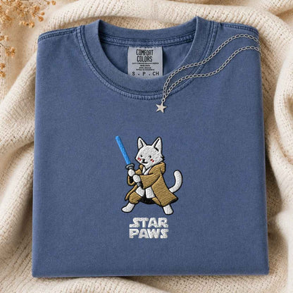 A folded slate blue t-shirt with a cute embroidered white cat character wearing a brown robe and holding a blue laser, with 'STAR PAWS' embroidered below. The shirt is styled on a beige knit blanket with a silver star necklace draped over the collar.