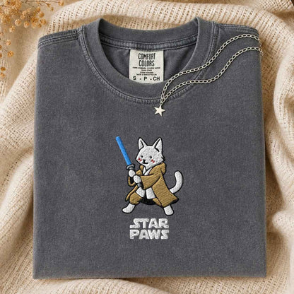 A folded dark gray t-shirt with a cute embroidered cat character wearing a brown robe and holding a blue laser, with the text 'STAR PAWS' below. The shirt is styled on a beige knit blanket with a silver star necklace draped over it.
