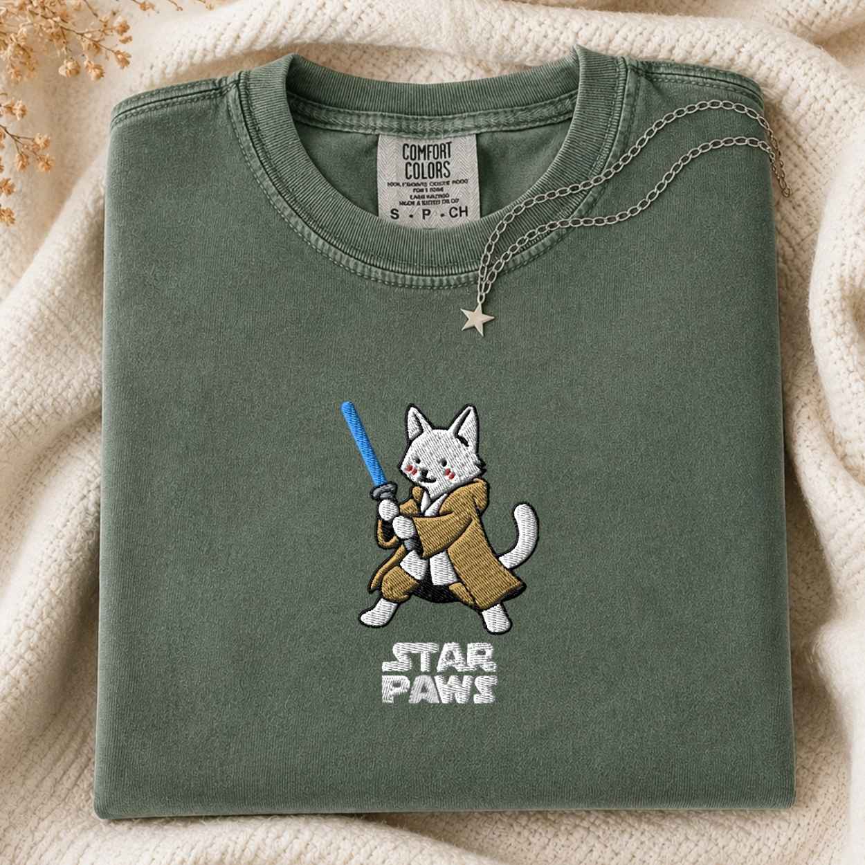 A folded sage green t-shirt with a cute embroidered cat character wearing a brown trench coat and holding a blue laser, with 'STAR PAWS' embroidered below. The shirt is styled on a cream knit blanket with a gold star necklace draped over the collar.