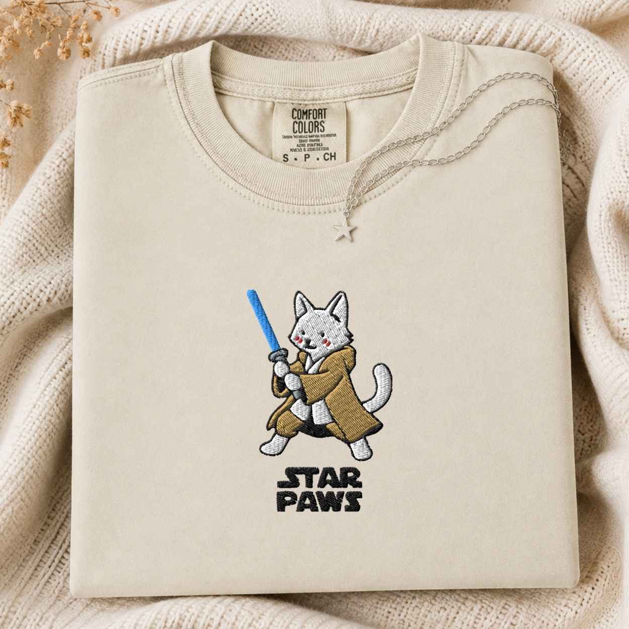 A folded cream-colored crewneck tshirt with a custom embroidered design of a white cat dressed as a protector holding a blue laser, above the text 'STAR PAWS'. The tshirt is laid flat on a cozy, textured cream knit blanket with dried flowers in the corner.