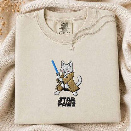 A folded cream-colored crewneck tshirt with a custom embroidered design of a white cat dressed as a protector holding a blue laser, above the text 'STAR PAWS'. The tshirt is laid flat on a cozy, textured cream knit blanket with dried flowers in the corner.