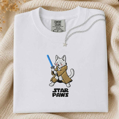 A folded white Comfort Colors t-shirt featuring a cartoon cat character holding a blue laser with the text 'STAR PAWS' underneath. The shirt is styled on a beige knit blanket with a silver necklace draped over the collar.