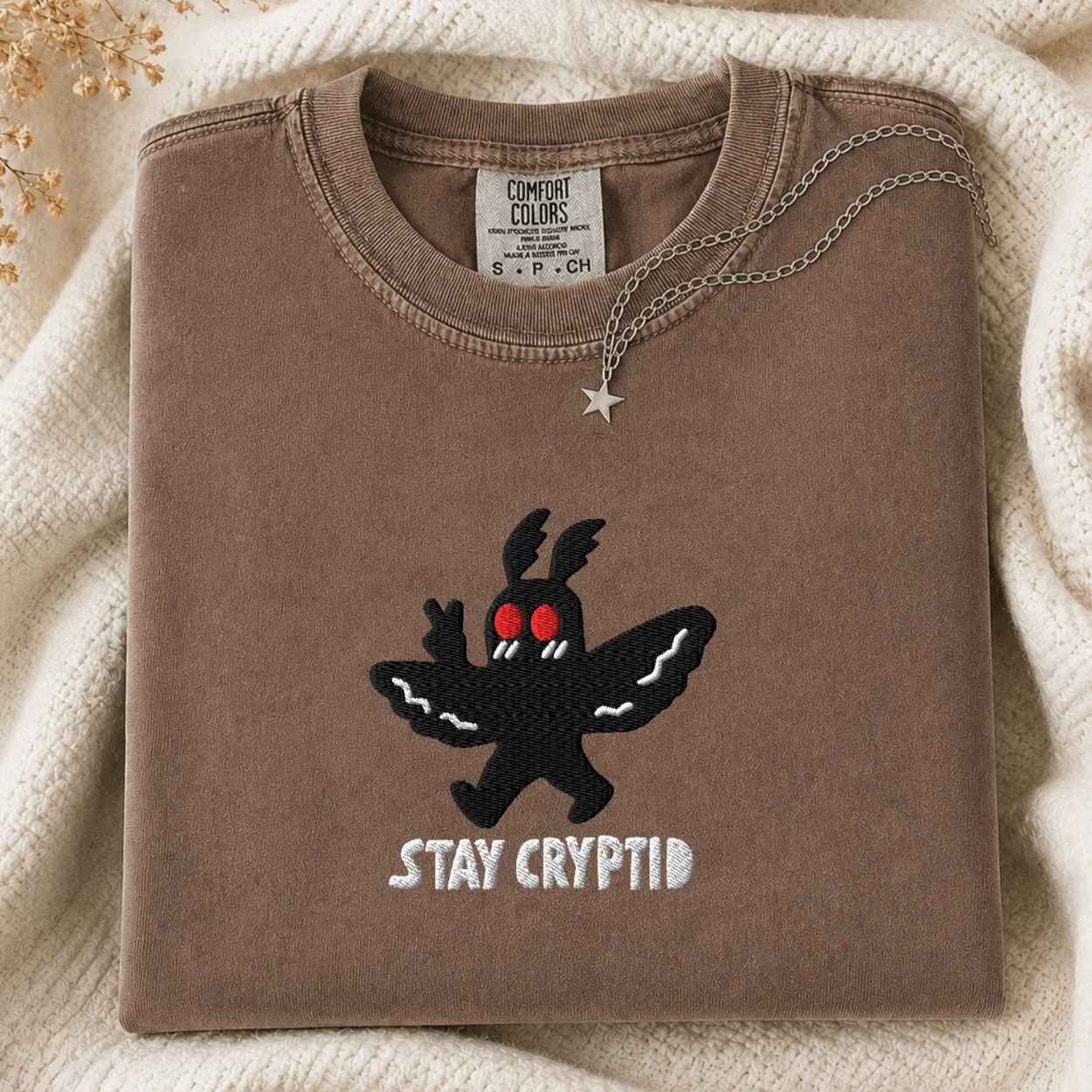 A folded brown sweatshirt with a black moth creature graphic and the words 'STAY CRYPTID' in white, styled on a cream knit blanket with a silver necklace draped over the collar. The inner neck label shows the brand 'Comfort Colors'.