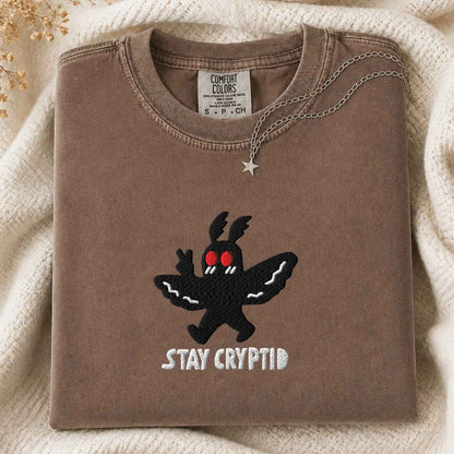 A folded brown sweatshirt with a black moth creature graphic and the words 'STAY CRYPTID' in white, styled on a cream knit blanket with a silver necklace draped over the collar. The inner neck label shows the brand 'Comfort Colors'.