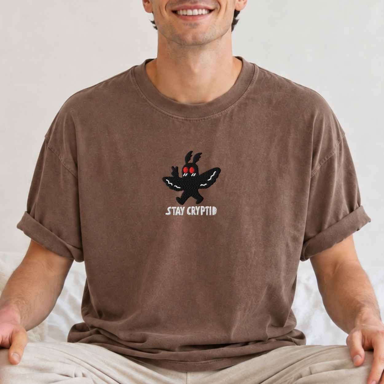 A man wearing a brown crewneck t-shirt with a cartoon bunny graphic and the text 'STAY CRYPTID' on the chest, paired with beige pants against a neutral light background.