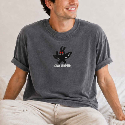 A model wearing gray t-shirt with a black embroidered cryptid character and the text 'STAY CRYPTID' in white, styled on a cream knit blanket with a silver necklace.