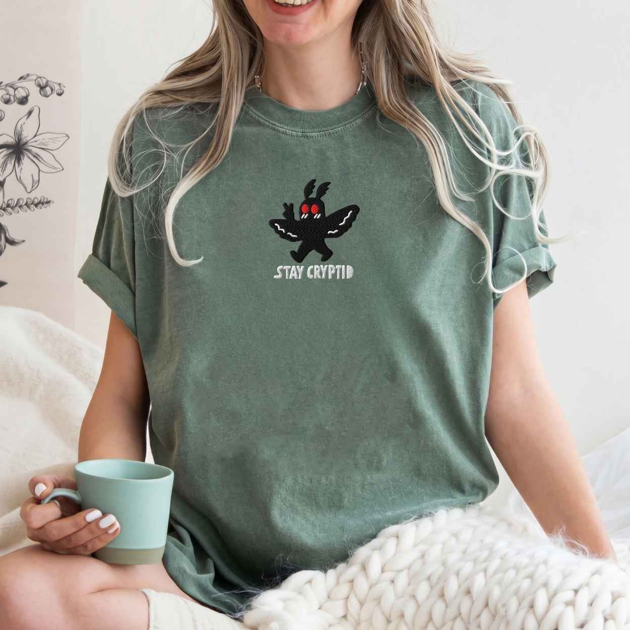 A model wearing sage green t-shirt with a black embroidered cryptid character and the text 'STAY CRYPTID' in white, styled on a cream knit blanket with a silver necklace.