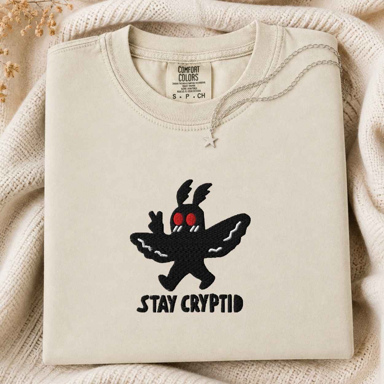 A folded cream-colored tshirt with a black embroidered Cryptid character and the text 'STAY CRYPTID' on the front, styled on a cozy cream knit blanket.