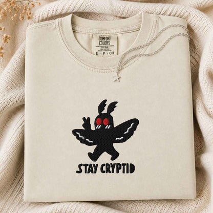 A folded cream-colored tshirt with a black embroidered Cryptid character and the text 'STAY CRYPTID' on the front, styled on a cozy cream knit blanket.