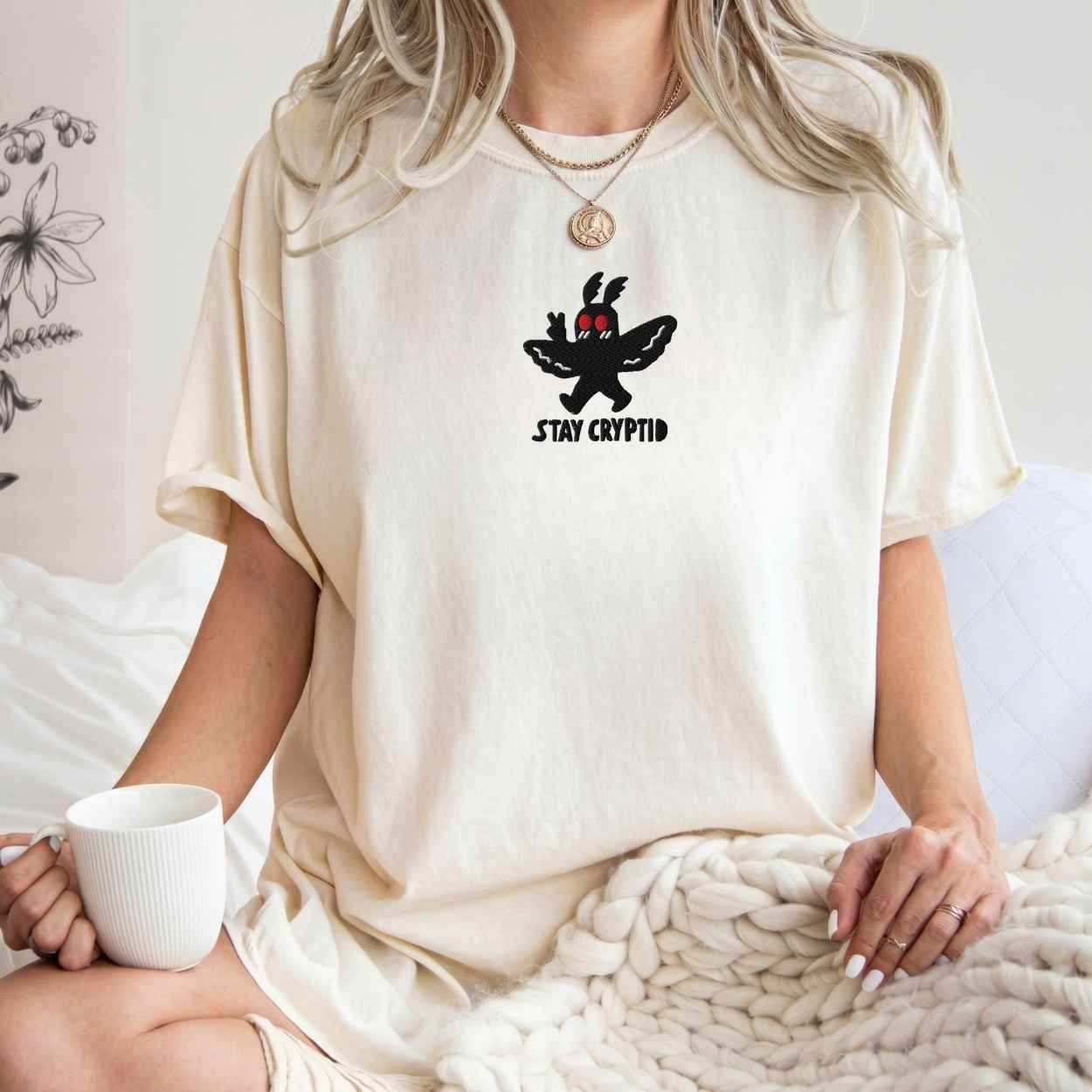 A model wearing white t-shirt with a black embroidered cryptid character and the text 'STAY CRYPTID' in black, styled on a cream knit blanket with a silver necklace.