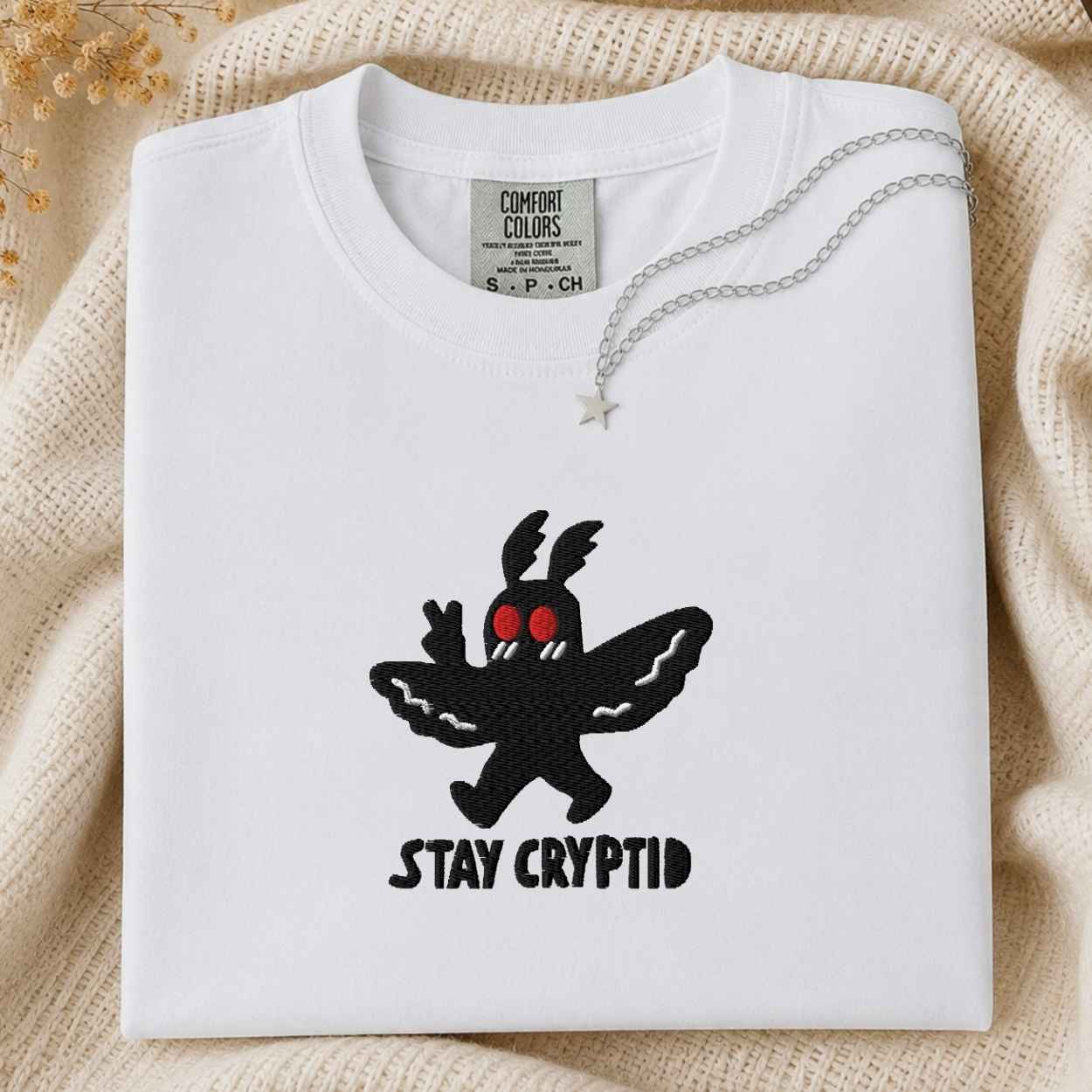 A folded sage white t-shirt with a black embroidered cryptid character and the text 'STAY CRYPTID' in black, styled on a cream knit blanket with a silver necklace.