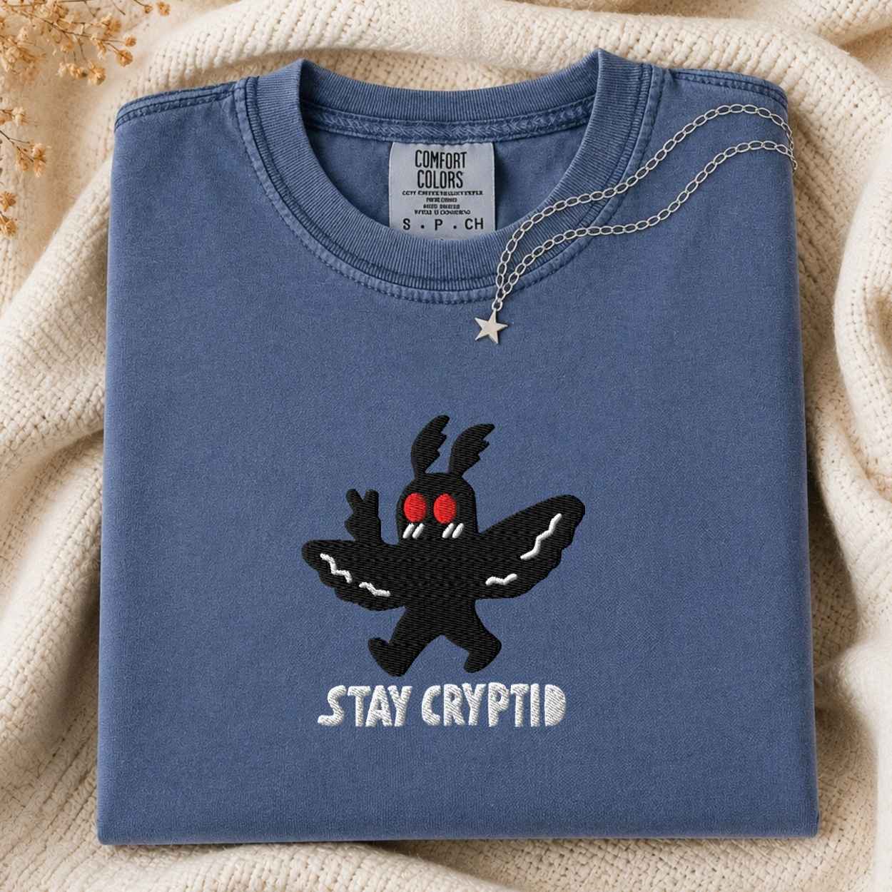 A folded slate blue crewneck tshirt with a black cryptid graphic and the words 'STAY CRYPTID' embroidered on the front, styled on a cream knit blanket with a silver necklace draped over the collar.