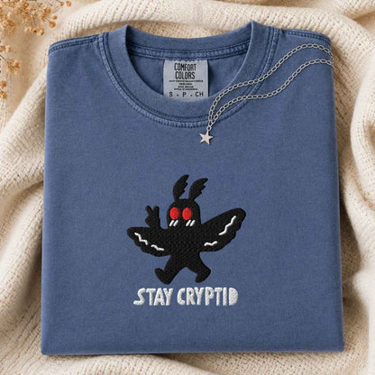 A folded slate blue crewneck tshirt with a black cryptid graphic and the words 'STAY CRYPTID' embroidered on the front, styled on a cream knit blanket with a silver necklace draped over the collar.
