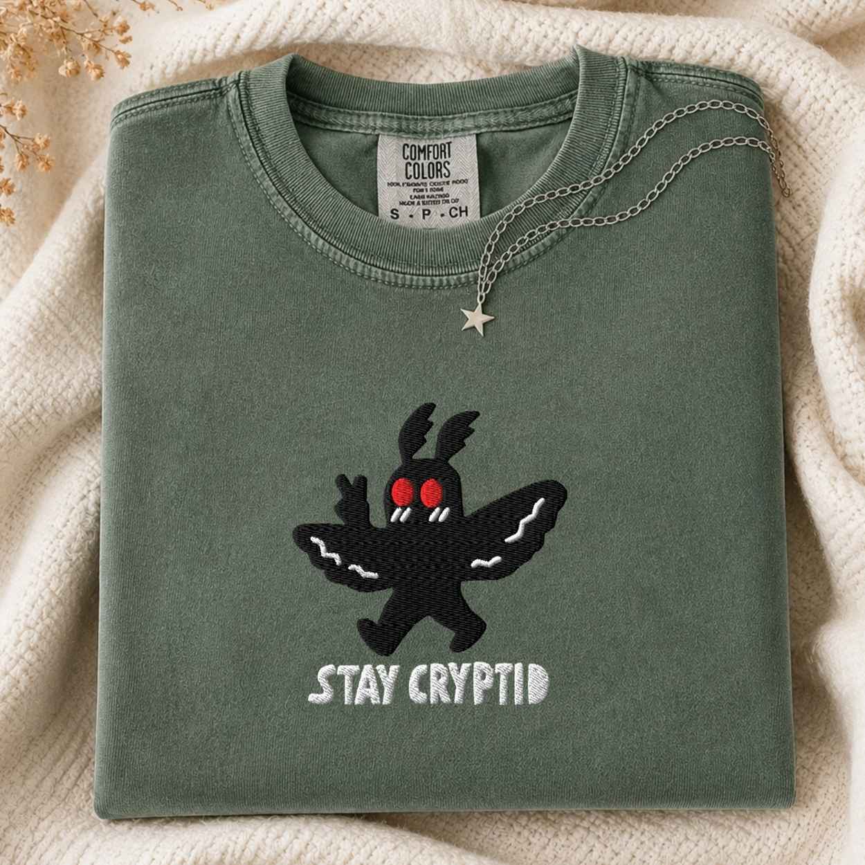 A folded sage green t-shirt with a black embroidered cryptid character and the text 'STAY CRYPTID' in white, styled on a cream knit blanket with a silver necklace.