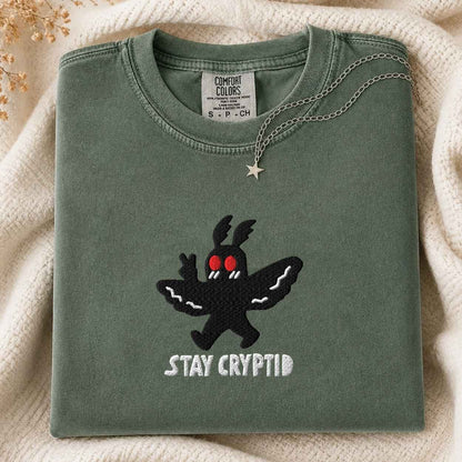 A folded sage green t-shirt with a black embroidered cryptid character and the text 'STAY CRYPTID' in white, styled on a cream knit blanket with a silver necklace.