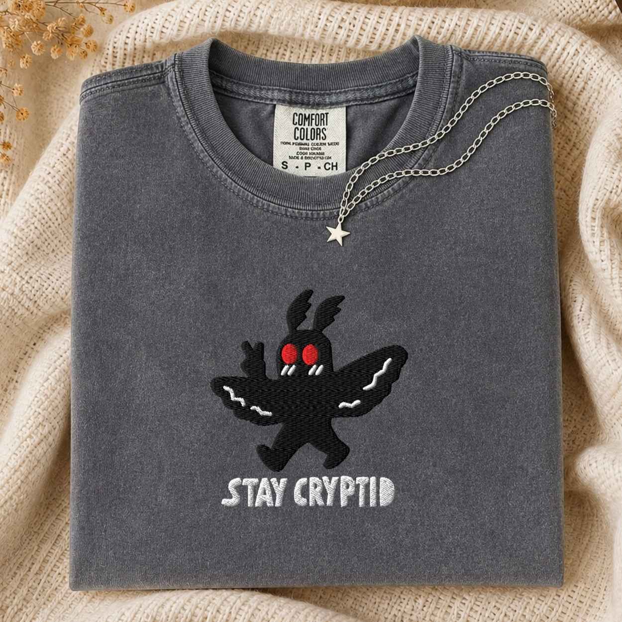 A folded sage gray t-shirt with a black embroidered cryptid character and the text 'STAY CRYPTID' in white, styled on a cream knit blanket with a silver necklace.