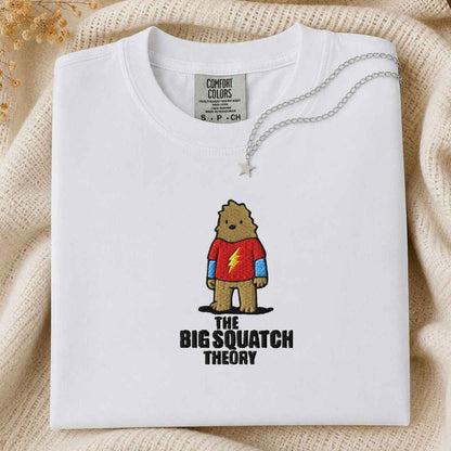 A folded white Comfort Colors t-shirt featuring a graphic of a cartoon bigfoot  in a red shirt with a yellow lightning bolt and the text 'THE BIG SQUATCH THEORY'. The shirt is styled on a beige knit blanket with a silver necklace draped over the collar.