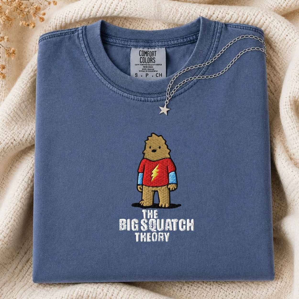 A folded slate blue t-shirt with a graphic of a cartoon bigfoot wearing a red shirt with a lightning bolt and the text 'THE BIG SQUATCH THEORY'. A silver star necklace is draped over the collar.
