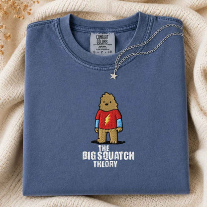 A folded slate blue t-shirt with a graphic of a cartoon bigfoot wearing a red shirt with a lightning bolt and the text 'THE BIG SQUATCH THEORY'. A silver star necklace is draped over the collar.