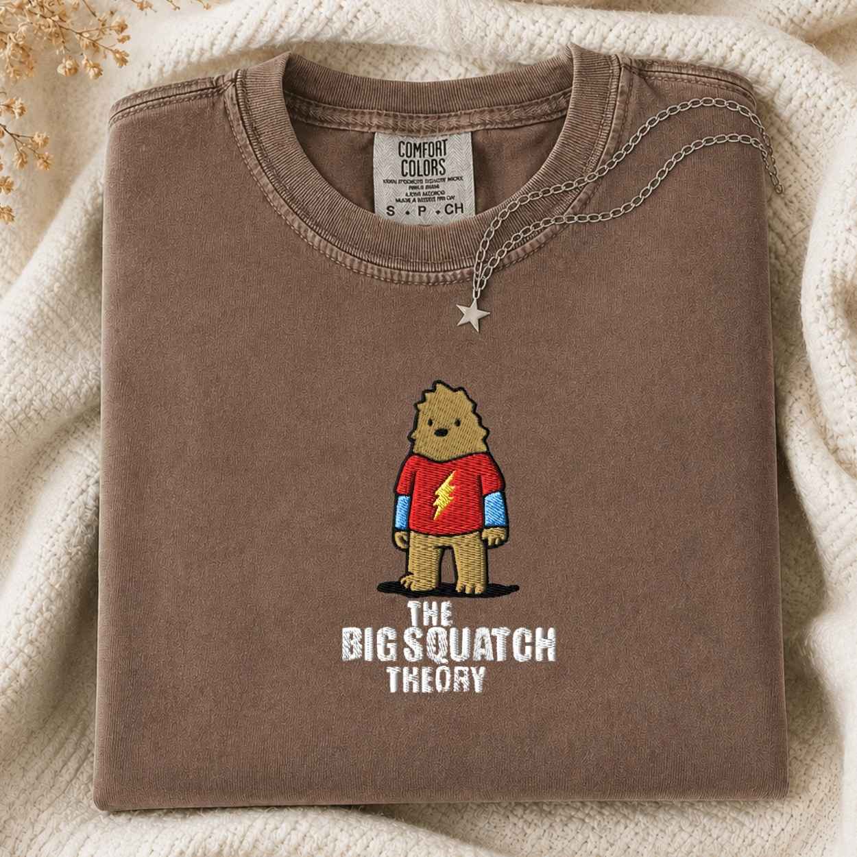A folded brown Comfort Colors t-shirt with a graphic of a cartoon bigfoot wearing a red shirt and the text 'THE BIG SQUATCH THEORY', styled on a cream knit blanket with a silver necklace.