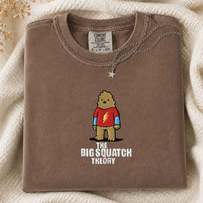 A folded brown Comfort Colors t-shirt with a graphic of a cartoon bigfoot wearing a red shirt and the text 'THE BIG SQUATCH THEORY', styled on a cream knit blanket with a silver necklace.