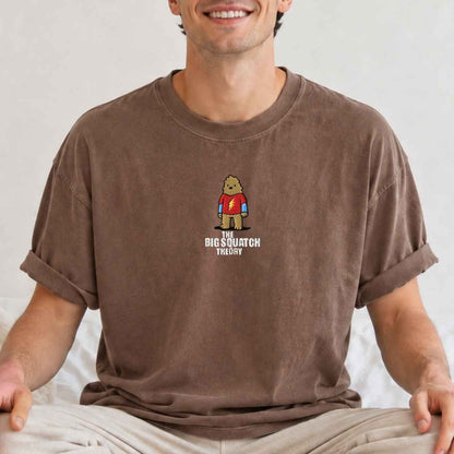A man wearing a brown crewneck t-shirt with a cartoon graphic of a bigfoot  wearing a red tshirt and the text 'The Big Squatch Theory' underneath.