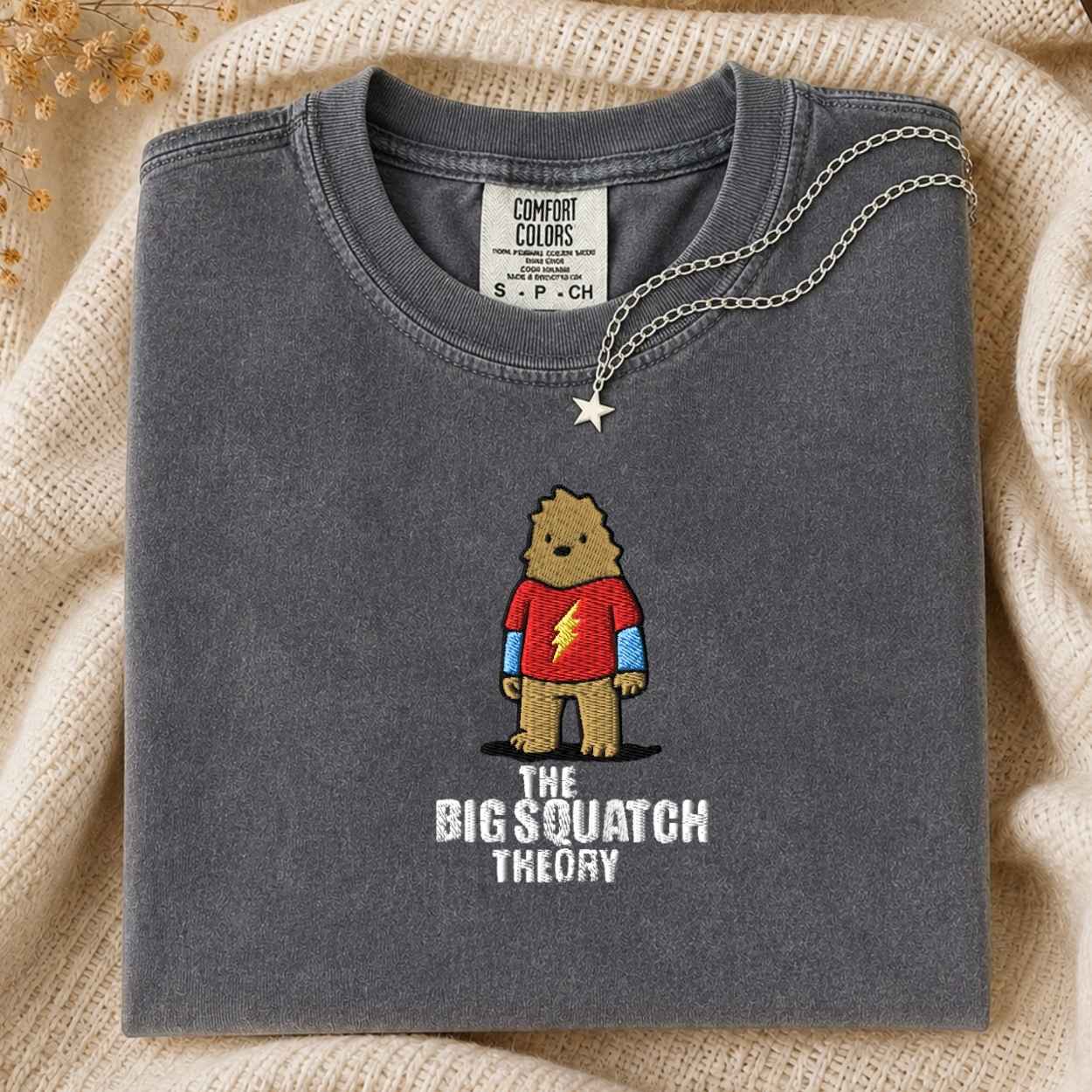 A folded dark gray Comfort Colors t-shirt featuring a cartoon graphic of a bigfoot in a red tshirt and blue full sleeve with the text 'THE BIG SQUATCH THEORY' underneath. A silver star pendant necklace is draped over the collar.