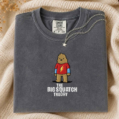 A folded dark gray Comfort Colors t-shirt featuring a cartoon graphic of a bigfoot in a red tshirt and blue full sleeve with the text 'THE BIG SQUATCH THEORY' underneath. A silver star pendant necklace is draped over the collar.
