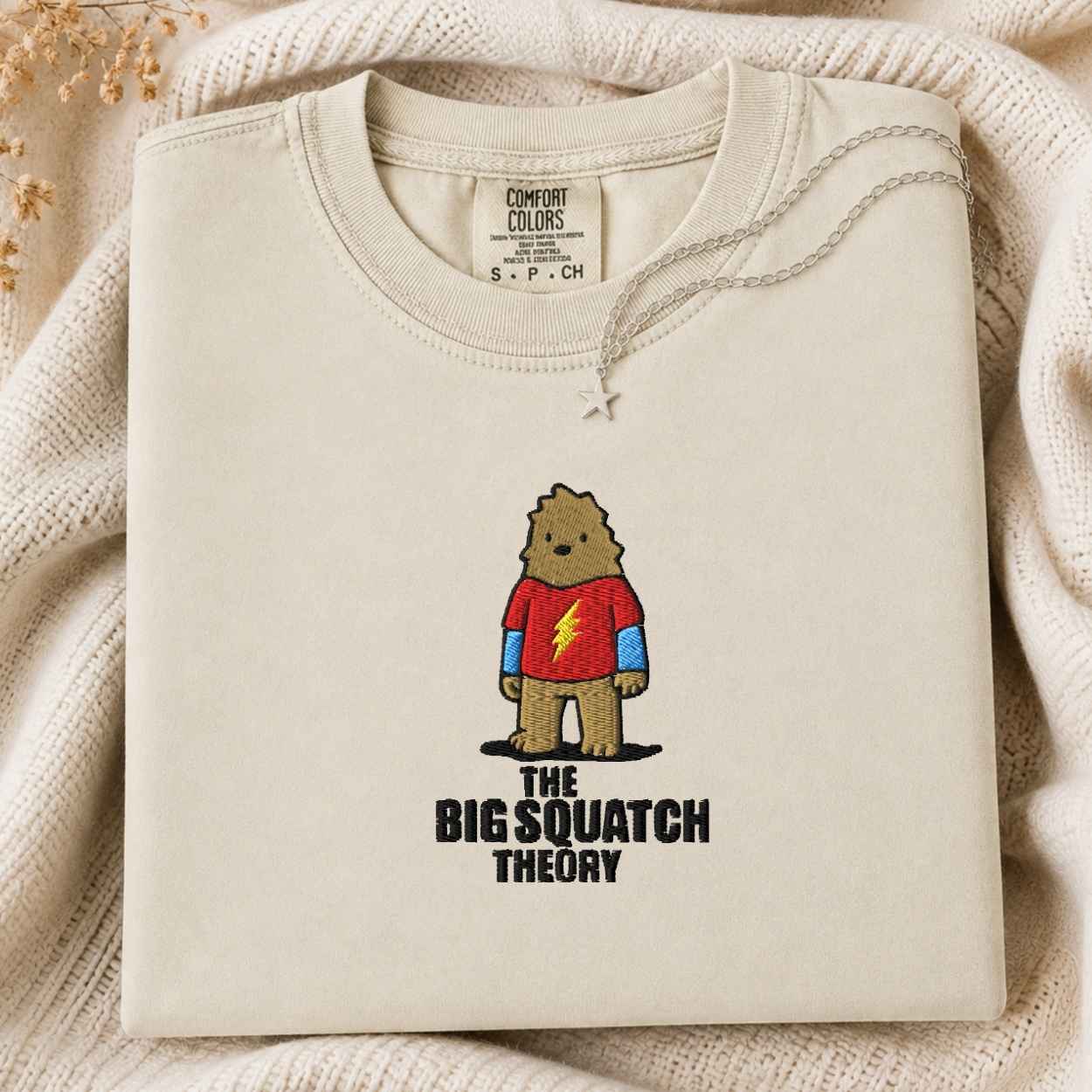 A folded cream-colored tshirt with a graphic of a bigfoot in a red shirt and the text 'THE BIG SQUATCH THEORY' underneath, styled on a textured cream knit blanket.