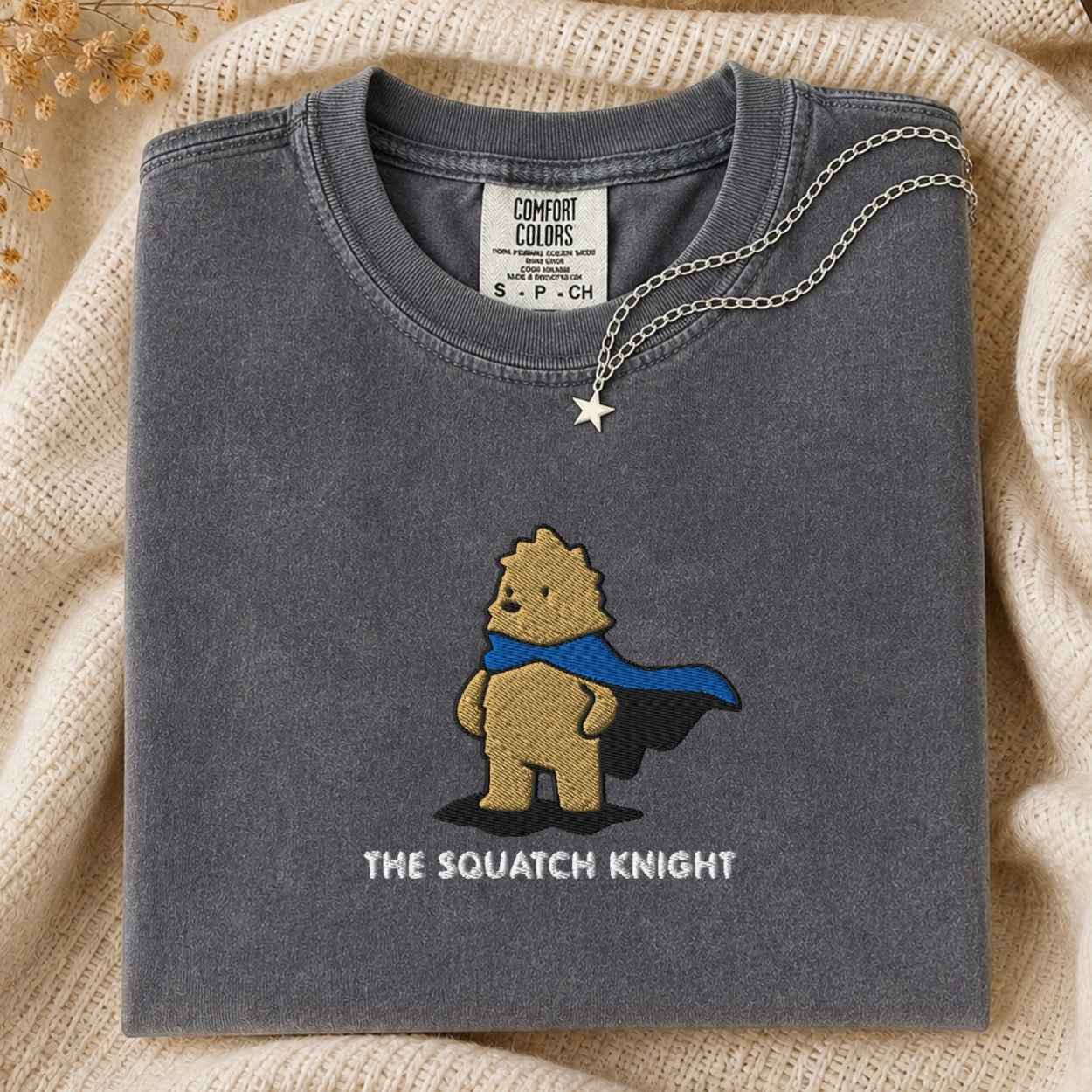 A folded dark gray t-shirt with a graphic of a bigfoot wearing a blue cape and the text 'THE SQUATCH KNIGHT' underneath. A silver star necklace is draped over the collar.