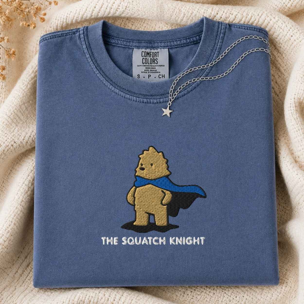 A folded slate blue t-shirt with a graphic of a bigfoot wearing a blue cape and the text 'THE SQUATCH KNIGHT' underneath. The shirt is styled on a cream-colored knit blanket with a silver necklace draped over the collar.