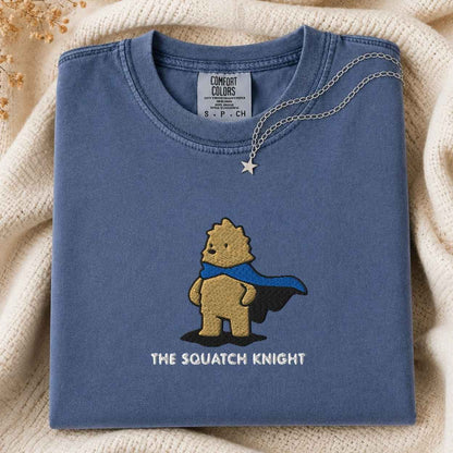 A folded slate blue t-shirt with a graphic of a bigfoot wearing a blue cape and the text 'THE SQUATCH KNIGHT' underneath. The shirt is styled on a cream-colored knit blanket with a silver necklace draped over the collar.