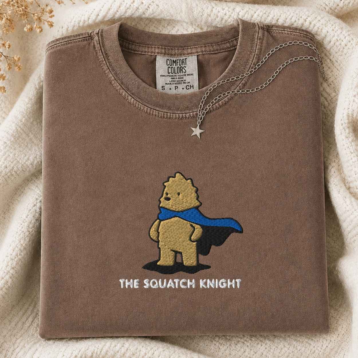 A folded brown tshirt with a graphic of a bigfoot wearing a blue cape and the text 'THE SQUATCH KNIGHT' underneath, styled on a cream knit blanket with a silver star necklace draped over the collar. The neck label reads 'COMFORT COLORS'.