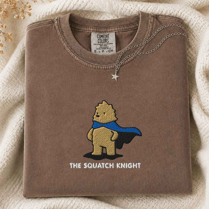 A folded brown tshirt with a graphic of a bigfoot wearing a blue cape and the text 'THE SQUATCH KNIGHT' underneath, styled on a cream knit blanket with a silver star necklace draped over the collar. The neck label reads 'COMFORT COLORS'.
