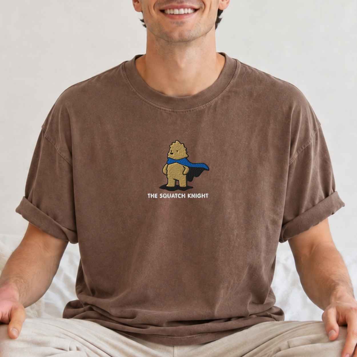 A man wearing a brown short-sleeve t-shirt with a cartoon graphic of a bigfoot in a blue cape labeled 'THE SQUATCH KNIGHT'.