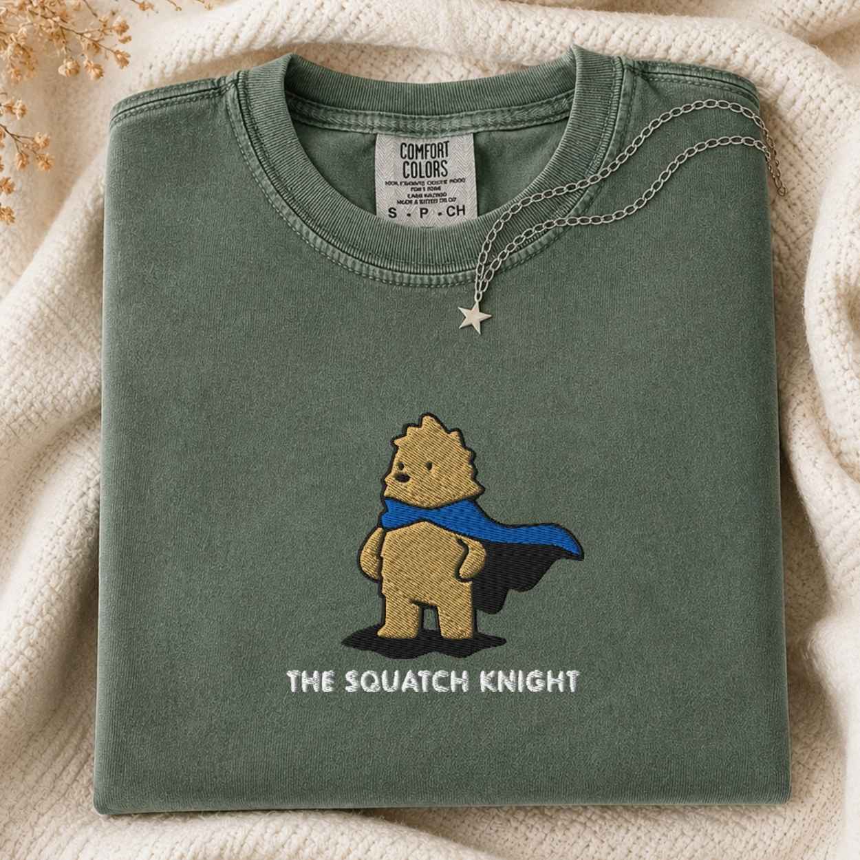 A folded sage green Comfort Colors t-shirt featuring an embroidered cartoon bigfoot superhero with a blue cape and the text 'THE SQUATCH KNIGHT'. The shirt is styled on a cream knit blanket with a silver star necklace draped over the collar.