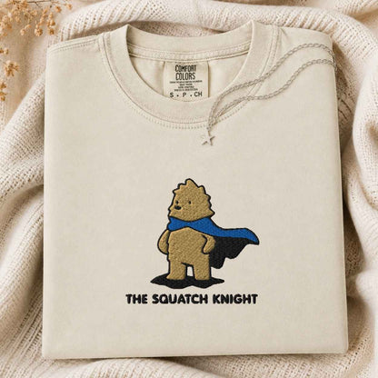 A folded cream-colored tshirt with a graphic of a bigfoot wearing a blue cape and the text 'THE SQUATCH KNIGHT' underneath, displayed on a cozy cream knit blanket.