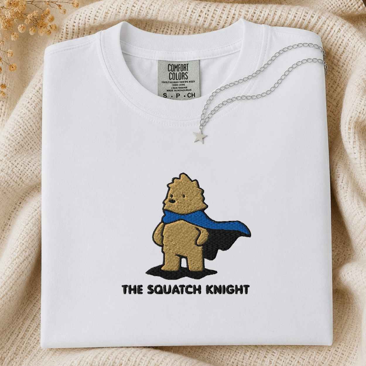 A folded white Comfort Colors t-shirt featuring an embroidered design of a bigfoot in a blue cape with the text 'THE SQUATCH KNIGHT' underneath. The shirt is styled on a beige knit blanket with a silver necklace draped over the collar.
