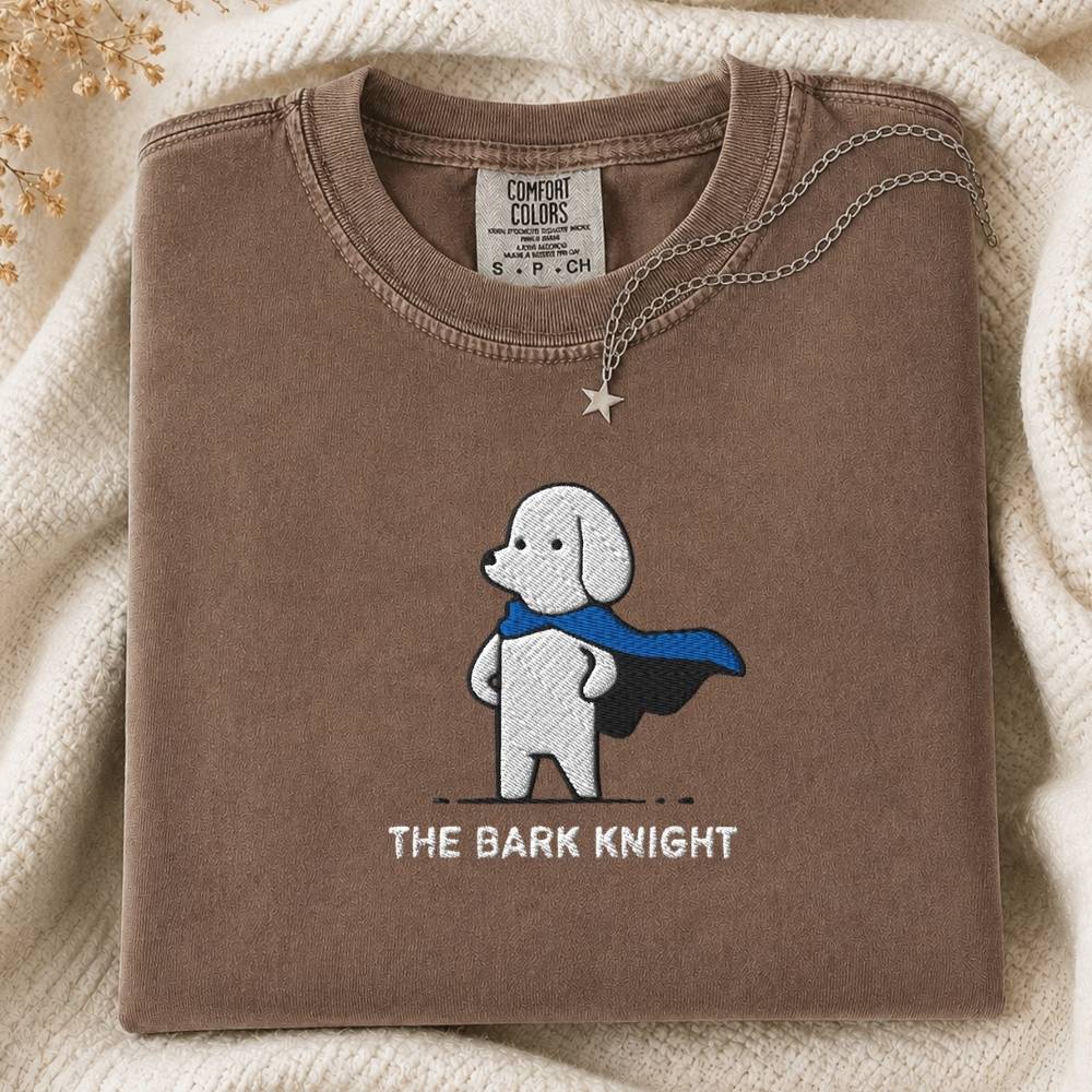 A folded brown t-shirt with a graphic of a white dog wearing a blue cape and the text 'THE BARK KNIGHT' underneath. The shirt is styled on a cream-colored knit blanket with a silver star pendant necklace draped over the collar.
