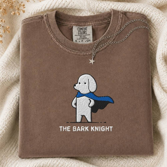 A folded brown t-shirt with a graphic of a white dog wearing a blue cape and the text 'THE BARK KNIGHT' underneath. The shirt is styled on a cream-colored knit blanket with a silver star pendant necklace draped over the collar.
