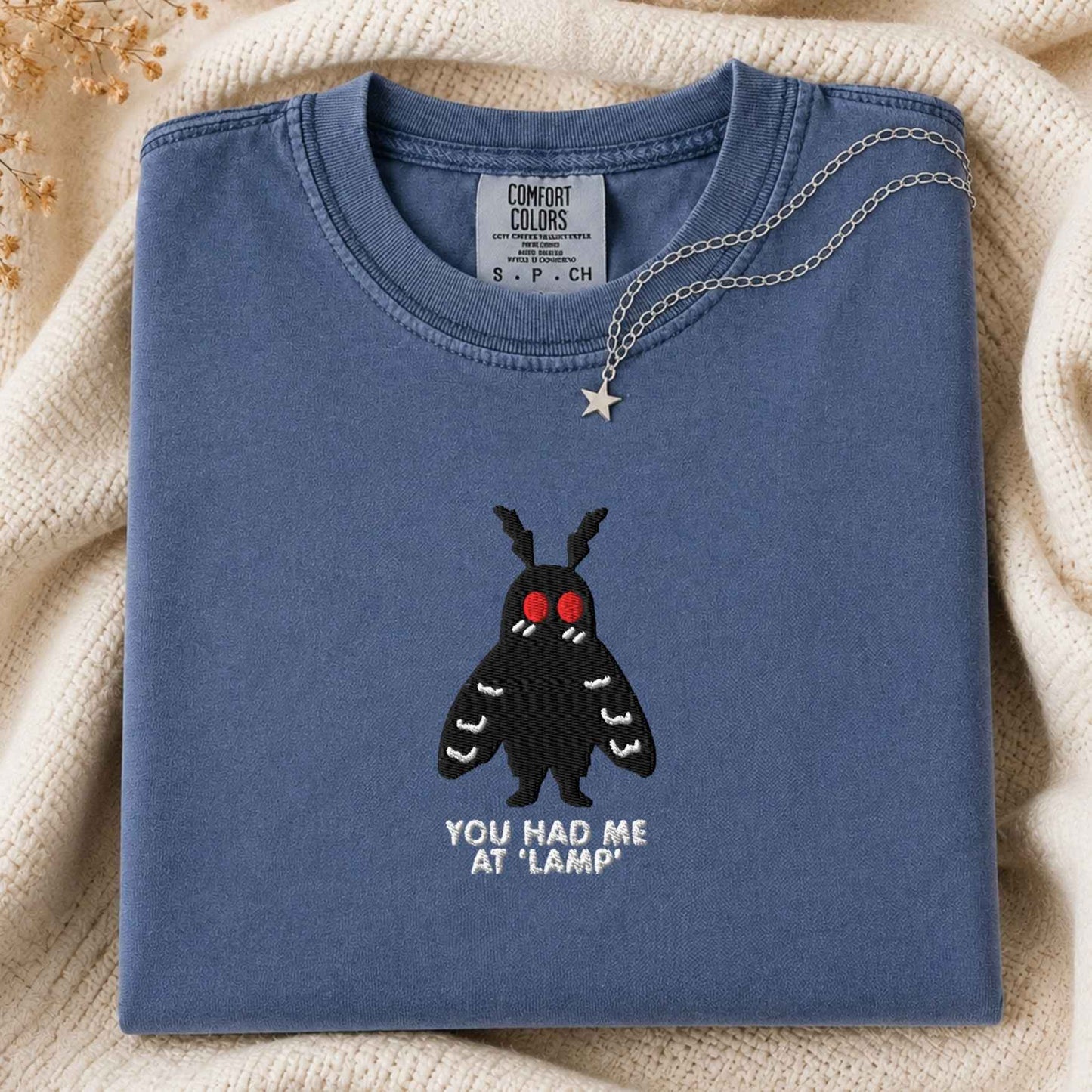 A folded slate blue t-shirt with a black moth graphic and the text 'YOU HAD ME AT "LAMP"' on the front, styled on a beige knit blanket with a silver necklace draped over the collar.