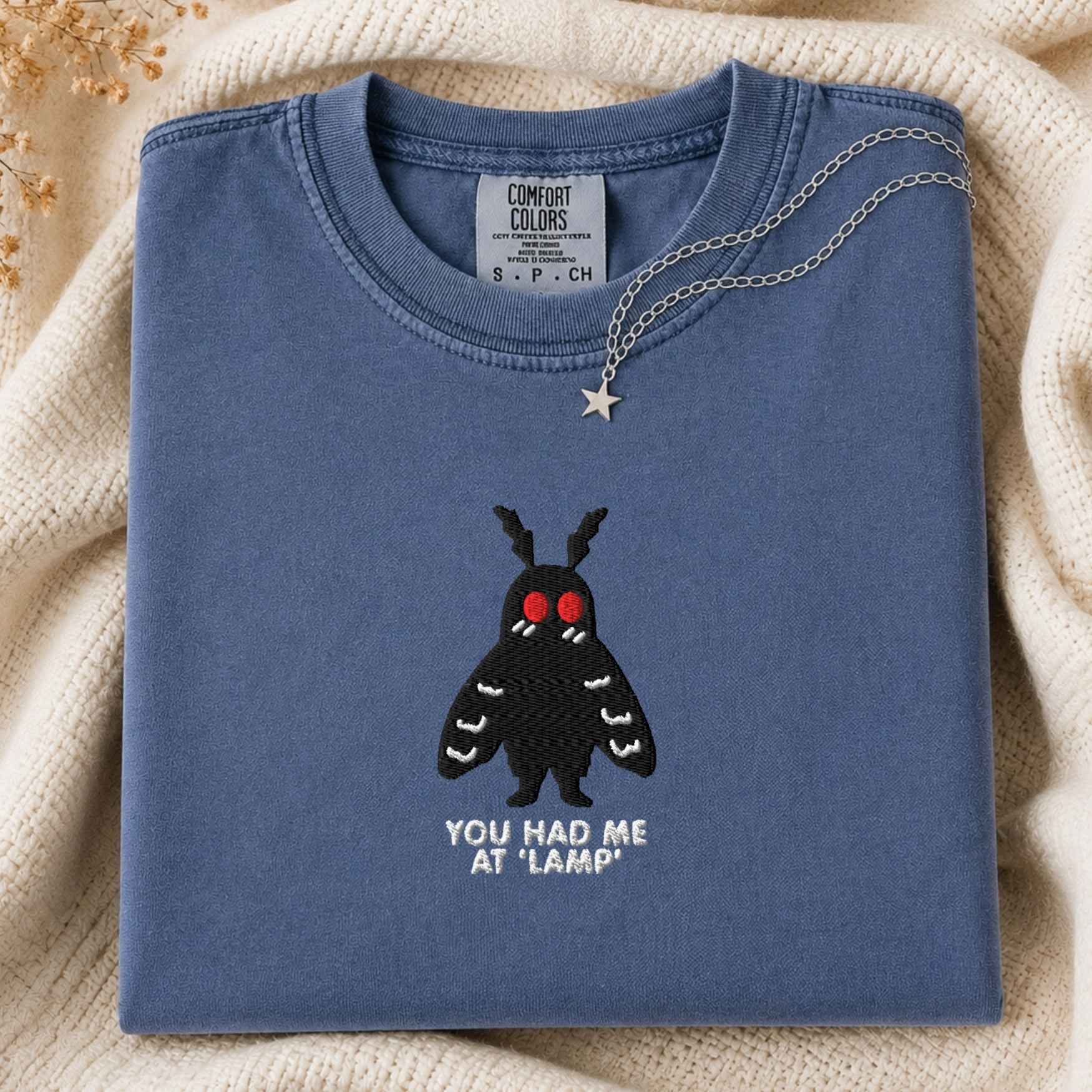 A folded slate blue t-shirt with a black moth graphic and the text 'YOU HAD ME AT "LAMP"' on the front, styled on a beige knit blanket with a silver necklace draped over the collar.