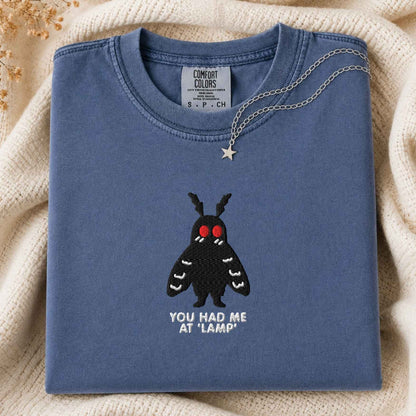 A folded slate blue t-shirt with a black moth graphic and the text 'YOU HAD ME AT "LAMP"' on the front, styled on a beige knit blanket with a silver necklace draped over the collar.